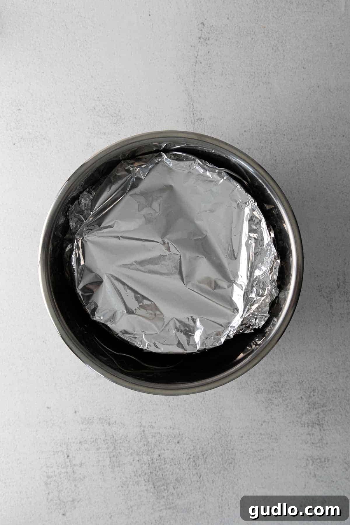 Cooking Your Cheesecake in the Instant Pot Foil-covered cheesecake in a springform pan being carefully lowered into an Instant Pot on a trivet.