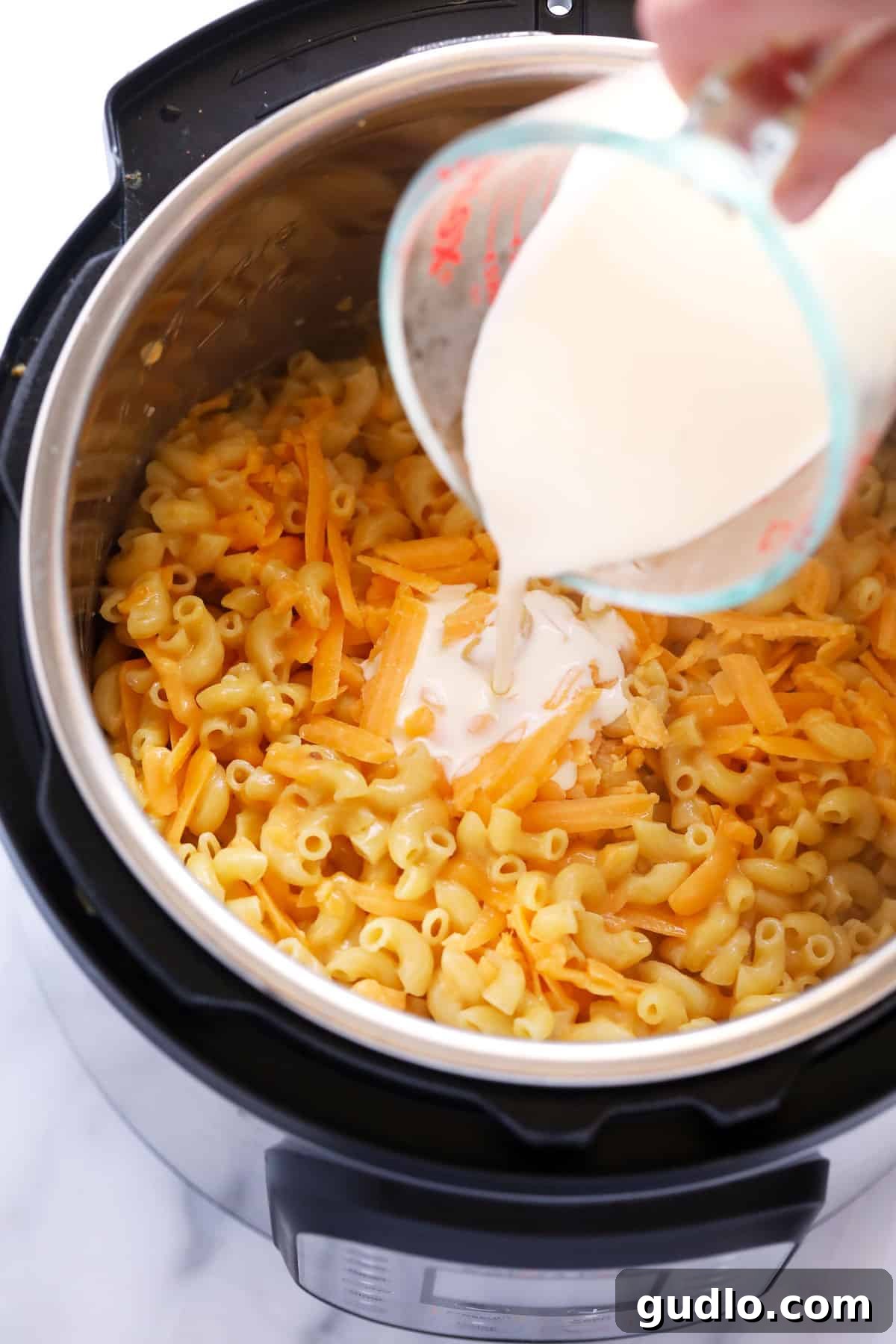 A hand pouring heavy cream into a steaming Instant Pot filled with cooked elbow macaroni and freshly melted cheddar cheese, creating a rich sauce.