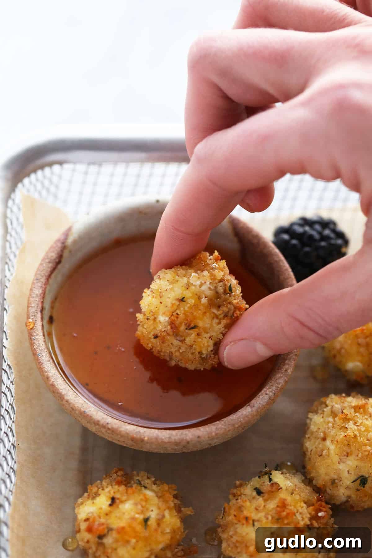 A hand dipping a crispy baked goat cheese ball into a small bowl of honey sriracha sauce.