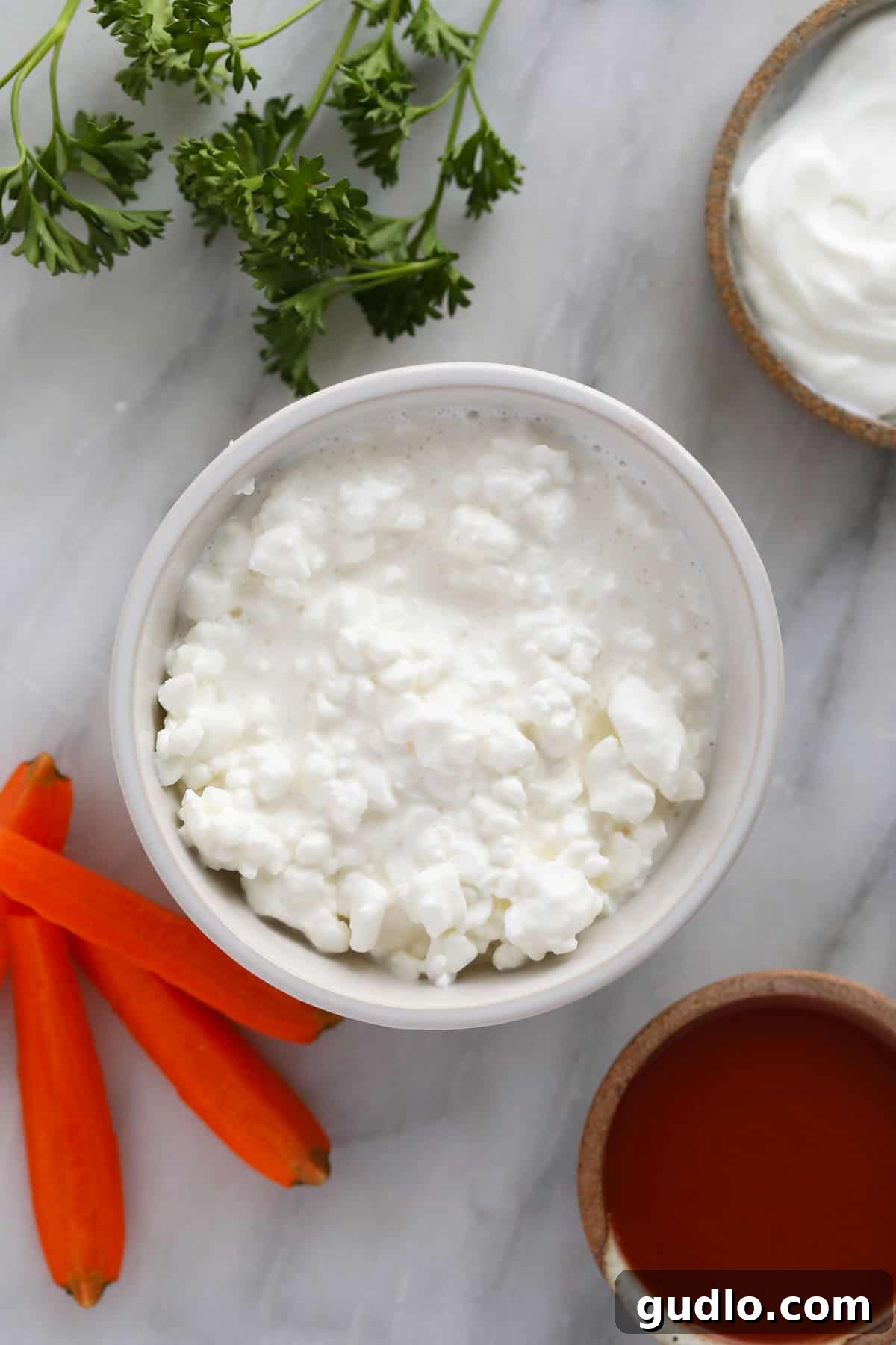 Essential Ingredients for Buffalo Whipped Cottage Cheese Dip Individual bowls containing full-fat cottage cheese, Greek yogurt, and Frank's RedHot buffalo sauce, arranged on a surface, signifying the core ingredients prepared for blending into a dip.