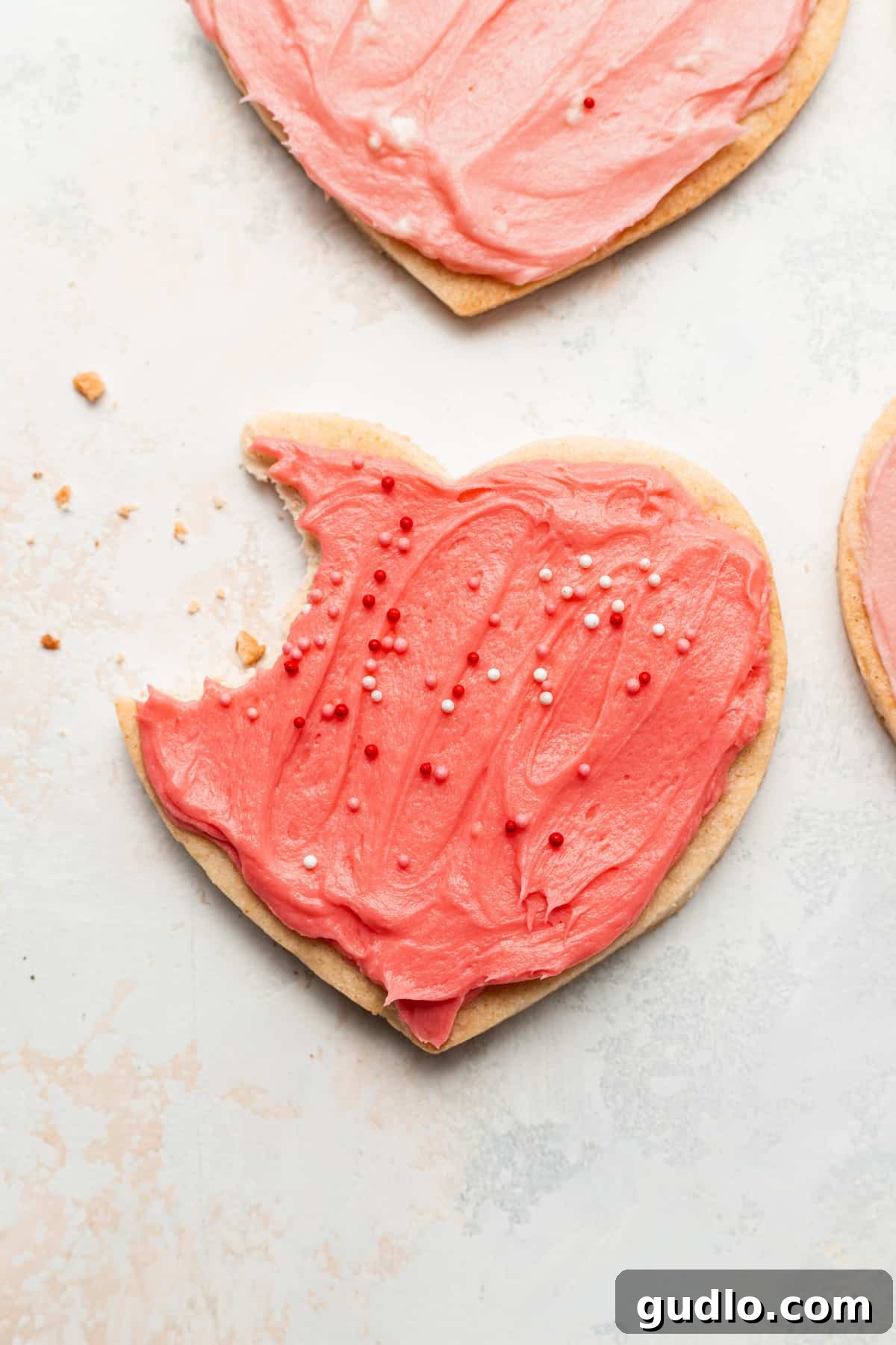 Yummy heart cookies for Valentine's Day with a homemade cream cheese frosting. These heart shaped cookies are so fun to bake as a family and made with our famous cream cheese sugar cookie dough. Enjoy! A perfectly frosted cream cheese heart cookie with pink frosting and sprinkles, a bite revealing its tender interior, symbolizing pure delight.