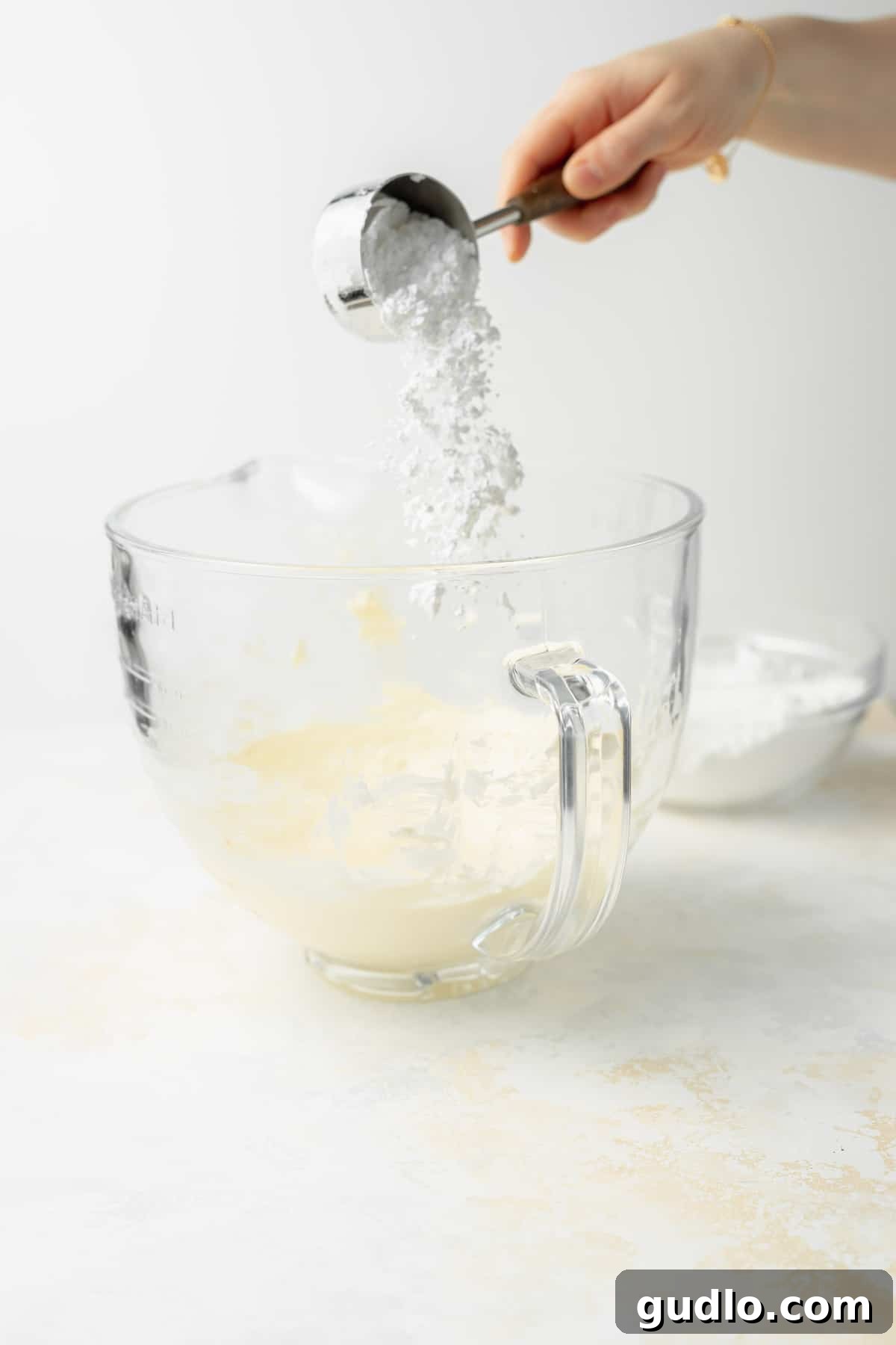 Adding Powdered Sugar for Smooth Cream Cheese Frosting A hand gently adding powdered sugar into a clear glass mixing bowl, capturing a key step in the frosting preparation.