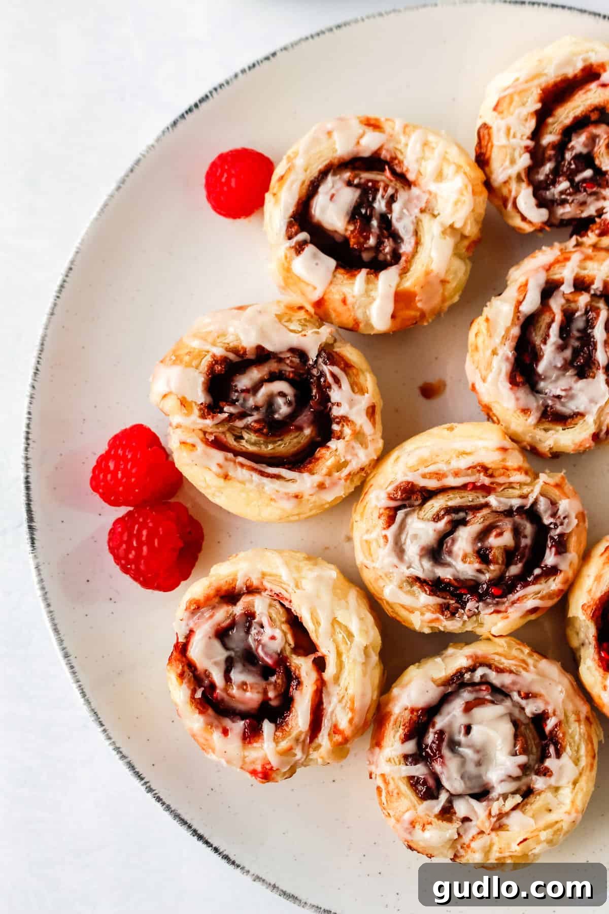 Start your day with these delicious Chocolate Raspberry Cinnamon Rolls that use store-bought dough and a few simple ingredients paired with a delicious cream cheese glaze. Freshly baked Chocolate Raspberry Cinnamon Rolls arranged elegantly on a white serving plate, generously drizzled with a creamy glaze and garnished with vibrant red raspberries, invitingly ready to be enjoyed as a luxurious breakfast or dessert.