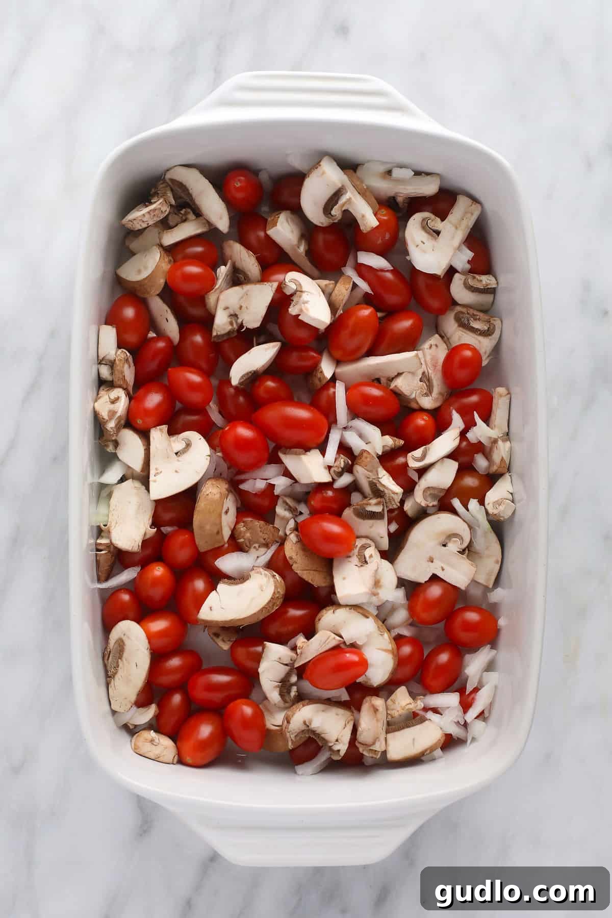 Ingredients Ready for Baking in a Casserole Dish Fresh ingredients for baked cream cheese pasta, including whole cream cheese block, cherry tomatoes, sliced mushrooms, and onion, artfully arranged in a casserole dish before baking.