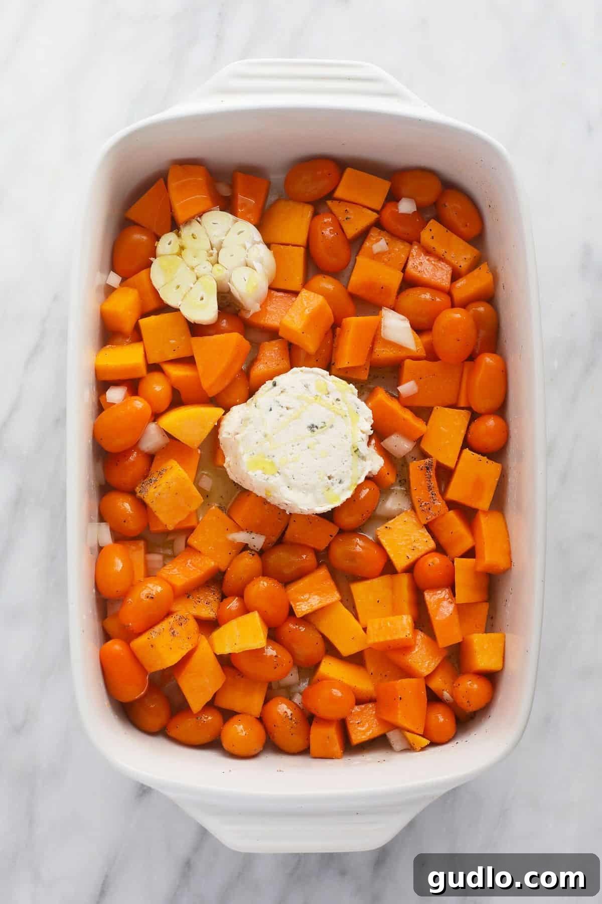 Ingredients Ready for Roasting Casserole dish with butternut squash, cherry tomatoes, onion, a whole head of garlic, and a puck of Boursin cheese before baking