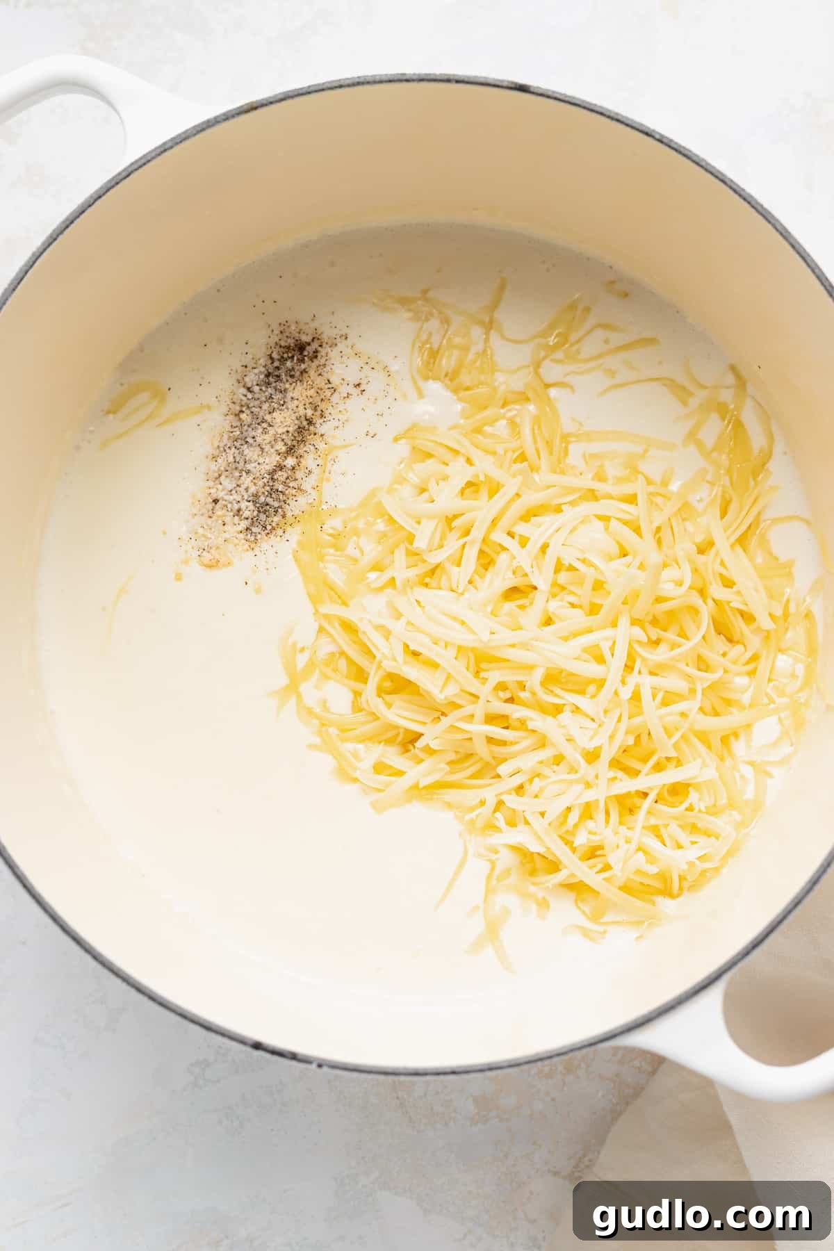 Adding shredded white cheddar cheese to the creamy roux in a pot on the stovetop.