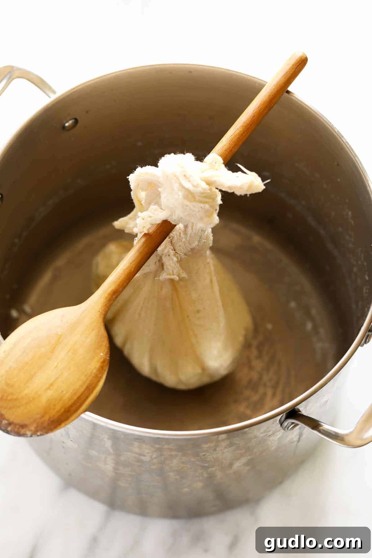 Crafting Fresh Paneer at Home 7 A ball of paneer wrapped in cheesecloth, hanging from a wooden spoon over a deep pot to drain.