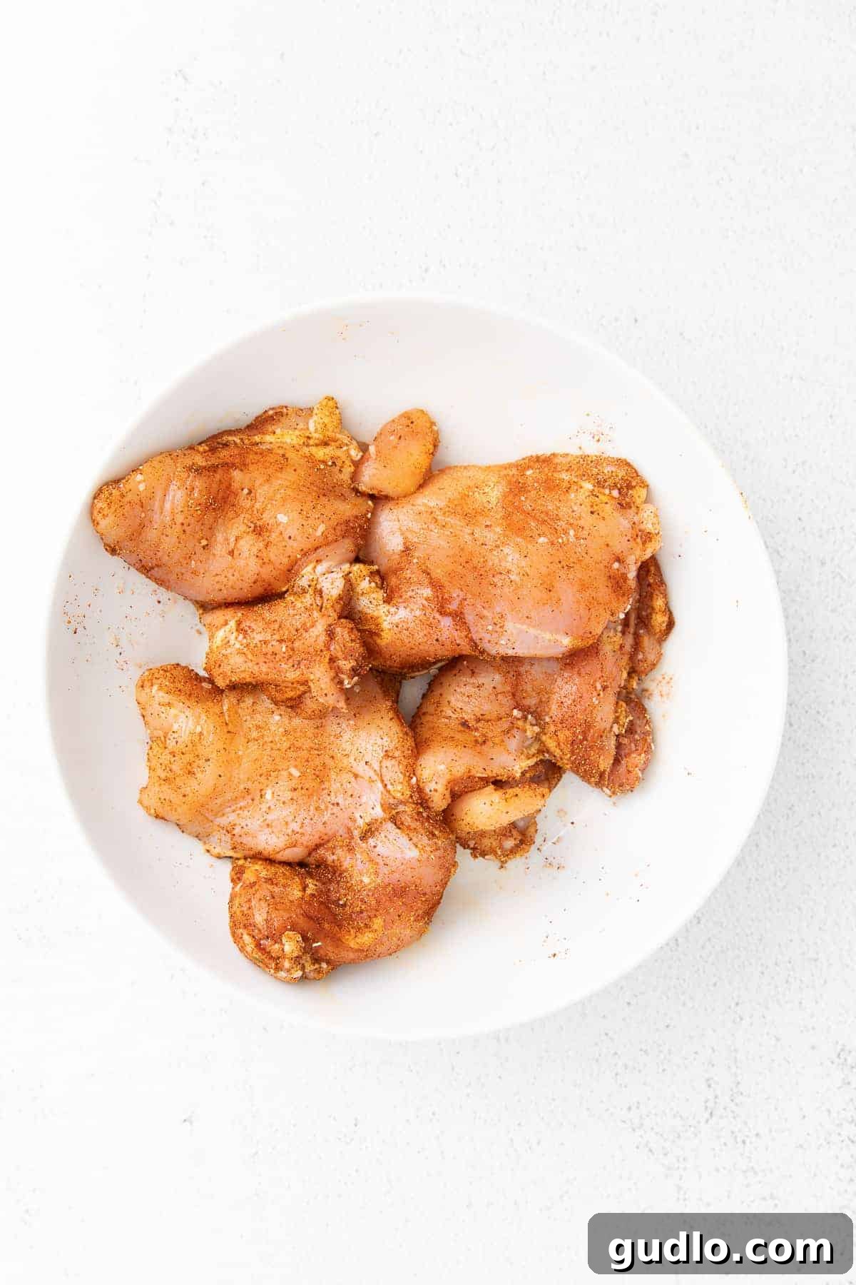 Marinated chicken thighs ready for cooking in a white bowl, seasoned with spices.
