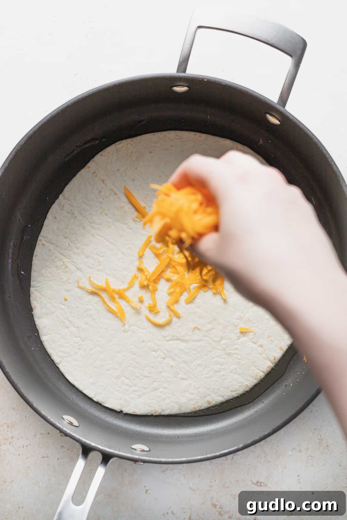 Sprinkling Cheese on a Tortilla in the Pan Hands sprinkling shredded cheddar cheese evenly onto a flour tortilla in a hot frying pan.