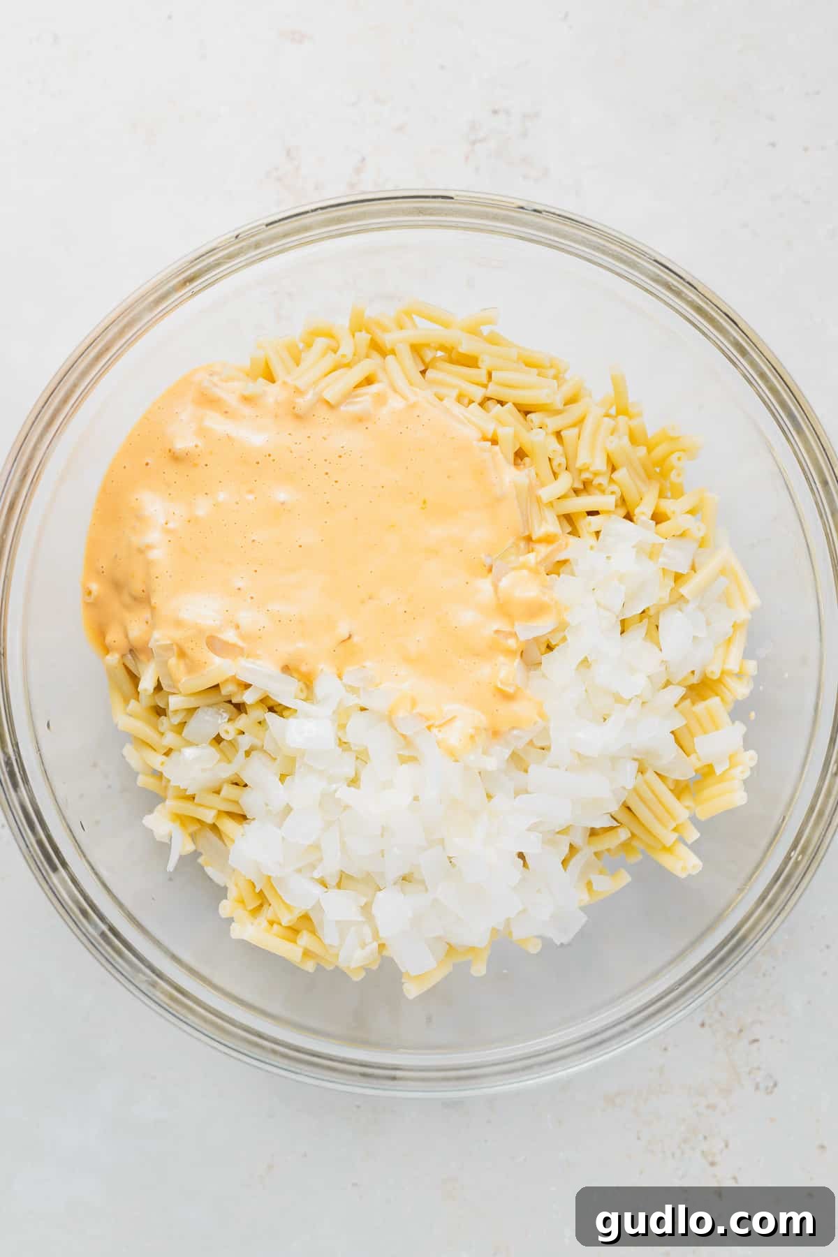 Combining the perfectly cooked macaroni with the rich, aromatic cheese sauce. Cooked macaroni noodles, creamy cheese sauce, and sautéed onion in a mixing bowl, ready to be combined.