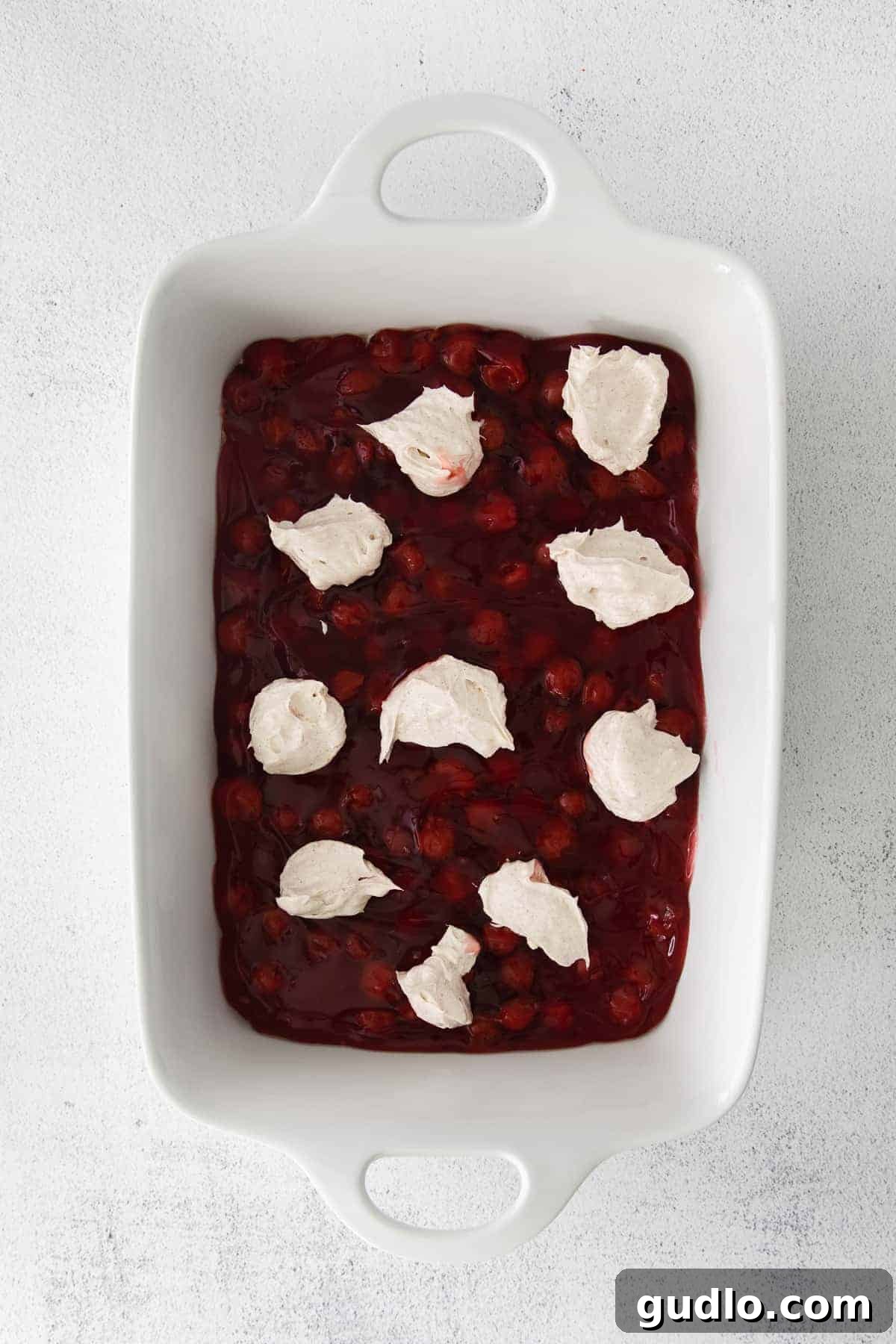 This Cream Cheese Cherry Dump Cake is calling your name! Dump all the ingredients into a cake pan, bake it, and enjoy topped with a scoop of vanilla ice cream. Unbaked cherry dump cake in a dish, showing layers of filling, cream cheese, and dry cake mix.