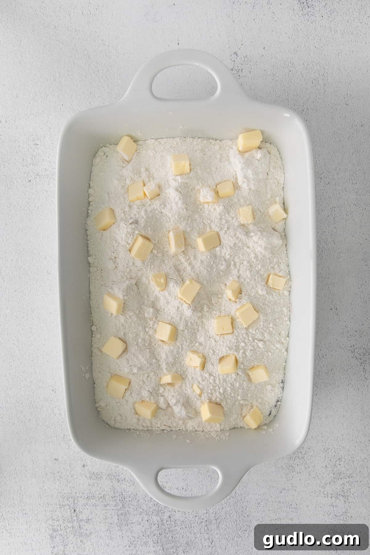 This Cream Cheese Cherry Dump Cake is calling your name! Dump all the ingredients into a cake pan, bake it, and enjoy topped with a scoop of vanilla ice cream. Raw dump cake in a casserole dish, topped with grated butter before baking.