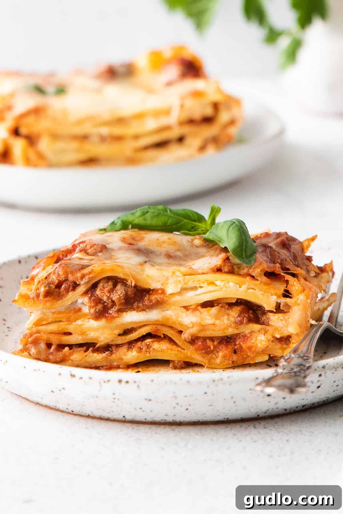 The Ultimate Meat Lasagna 2 A delicious slice of meat lasagna served on a white plate with a fork, garnished with fresh basil.