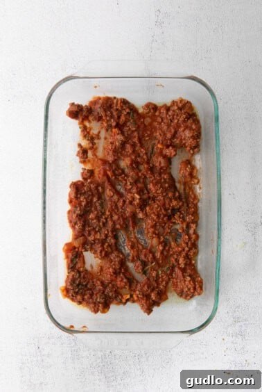 A glass baking dish with a base layer of rich meat sauce spread evenly, ready for the first layer of lasagna noodles.