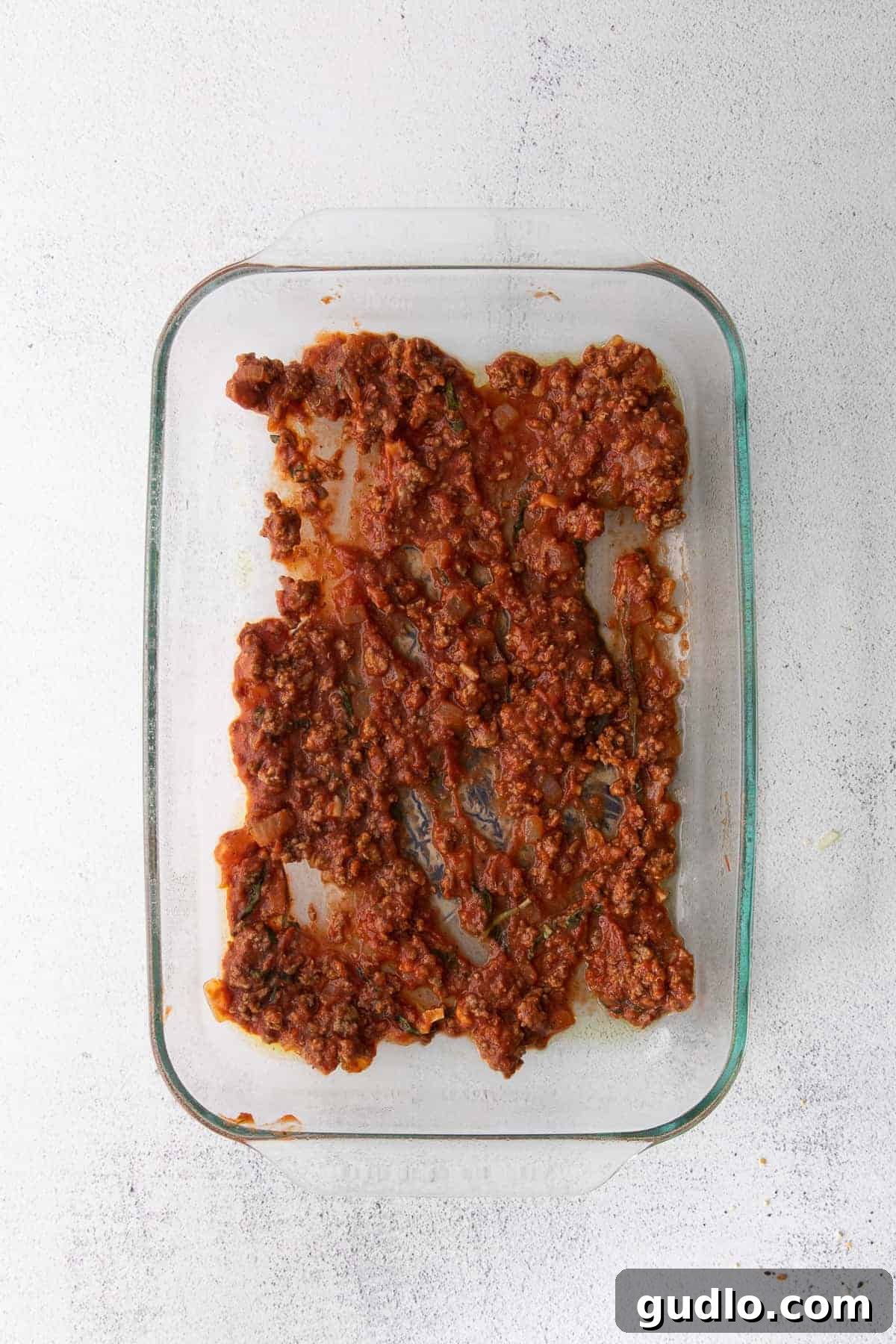 The Ultimate Meat Lasagna 4 A glass baking dish with a base layer of rich meat sauce spread evenly at the bottom, ready for layering lasagna noodles.
