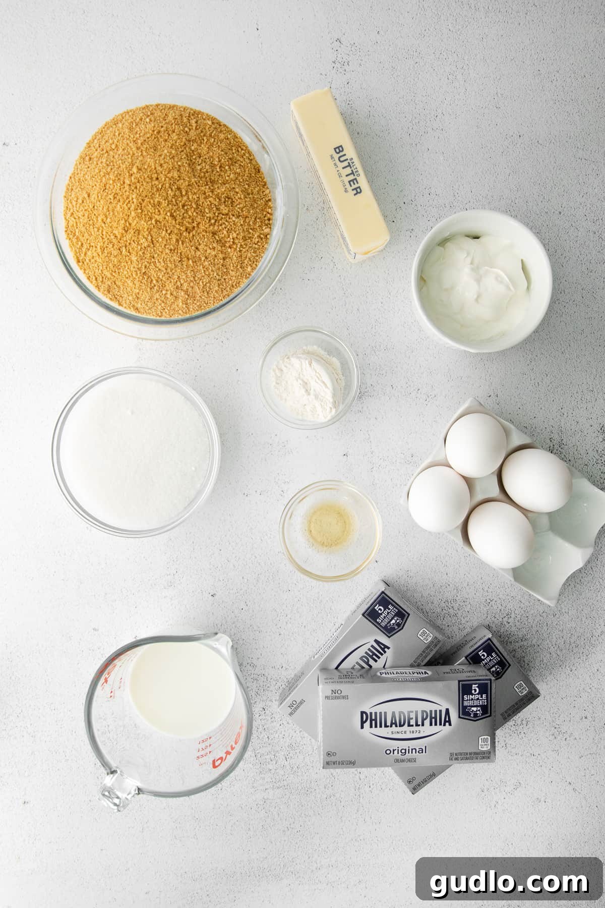 Individual bowls containing cheesecake ingredients like cream cheese, sugar, eggs, and vanilla, arranged on a kitchen counter.