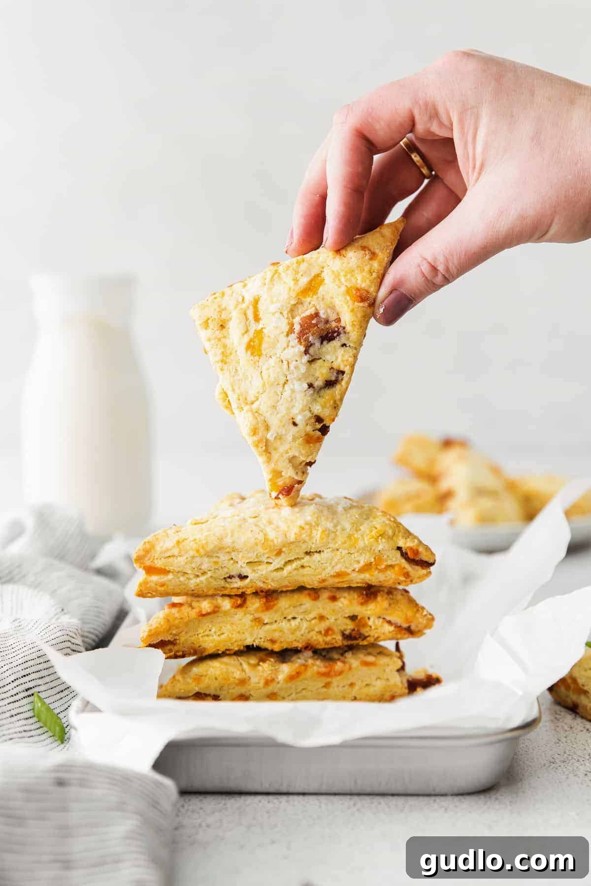A hand holding a golden-brown bacon and cheddar scone, showing its flaky texture and savory fillings.