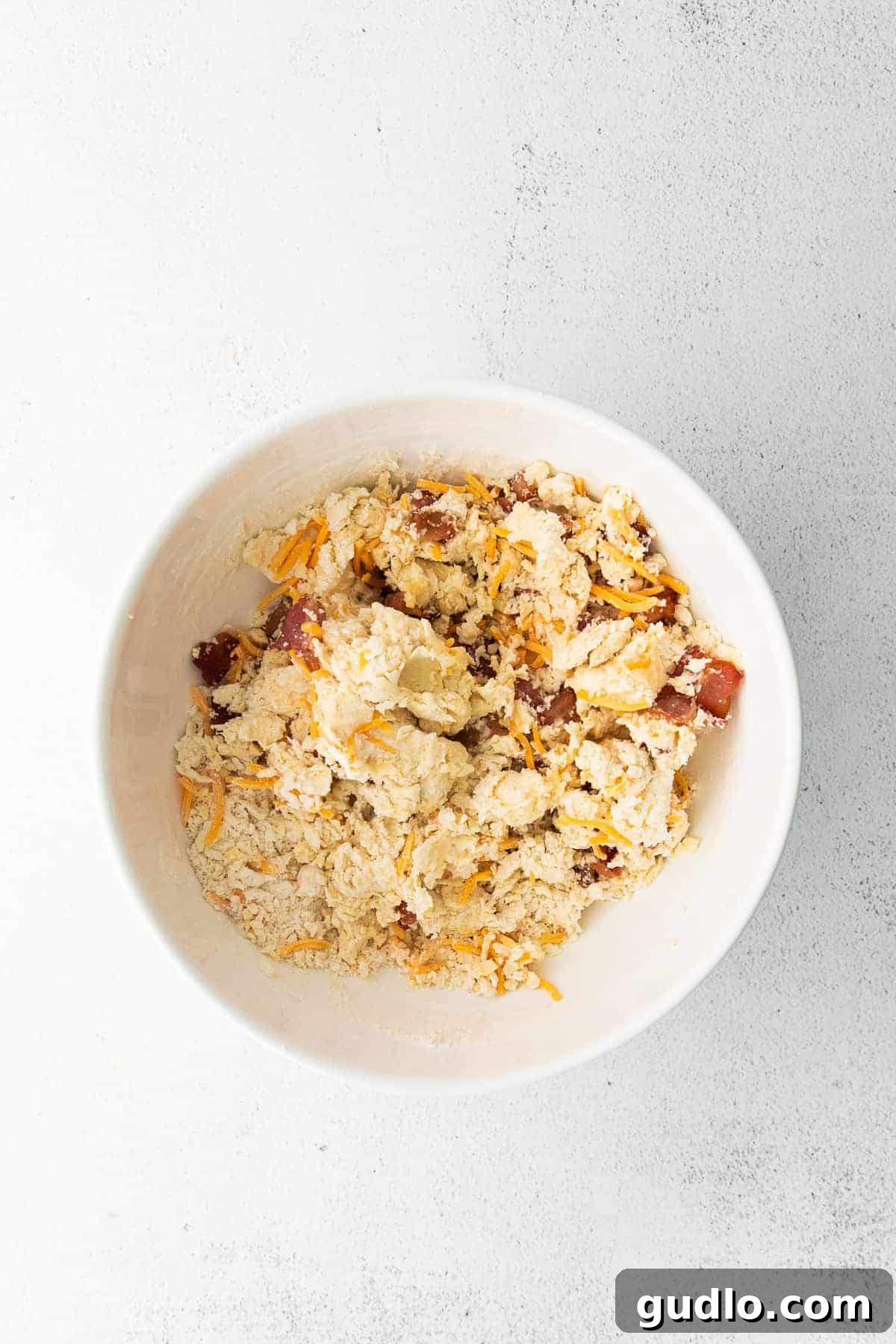 A large bowl filled with crumbly bacon and cheddar scone dough, ready to be shaped and baked.