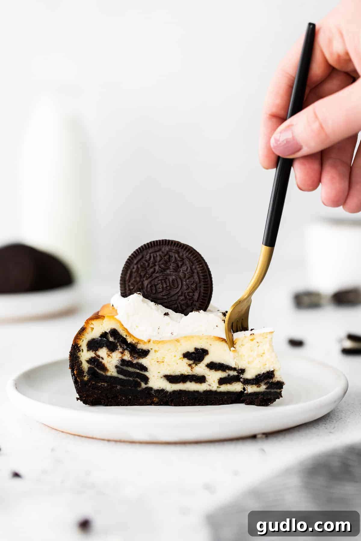 A close-up of a slice of Oreo cheesecake on a white plate, with a fork taking a bite, showing the creamy filling and chunky Oreo pieces.