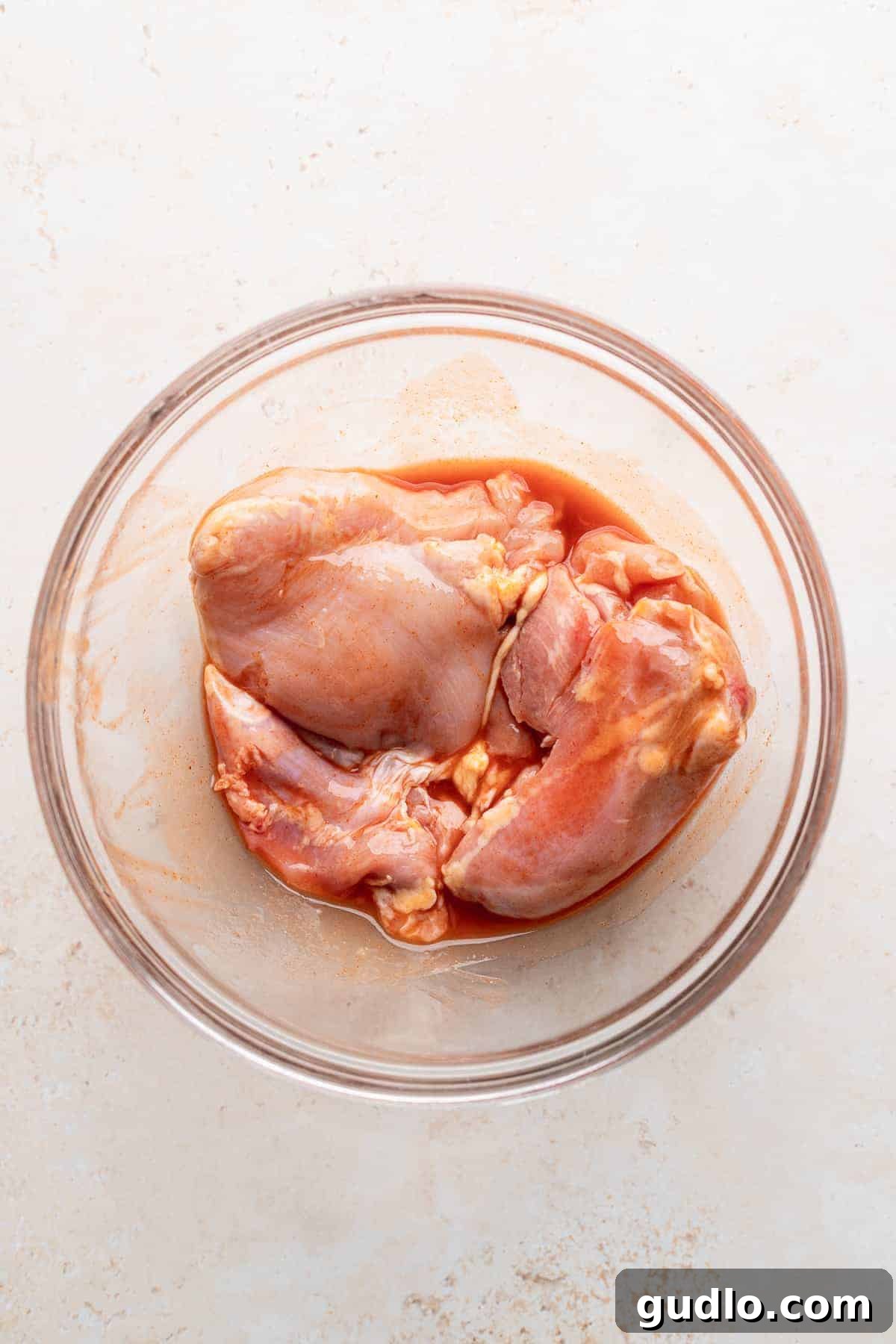 Boneless, skinless chicken thighs marinating in a vibrant red buffalo hot sauce and honey mixture in a clear glass bowl, resting on a clean white surface, showcasing the preparation process.