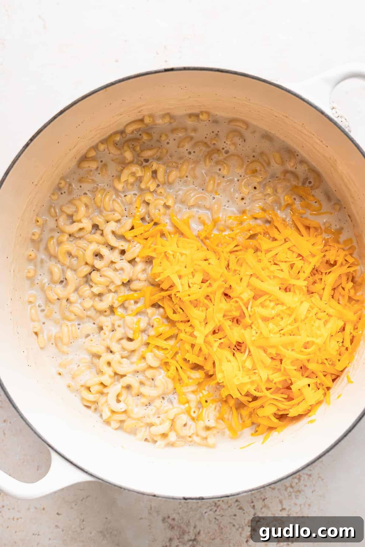 Close-up of freshly shredded yellow cheddar cheese sprinkled generously over cooked macaroni noodles in a large pot, ready to be stirred into a creamy, melting sauce.
