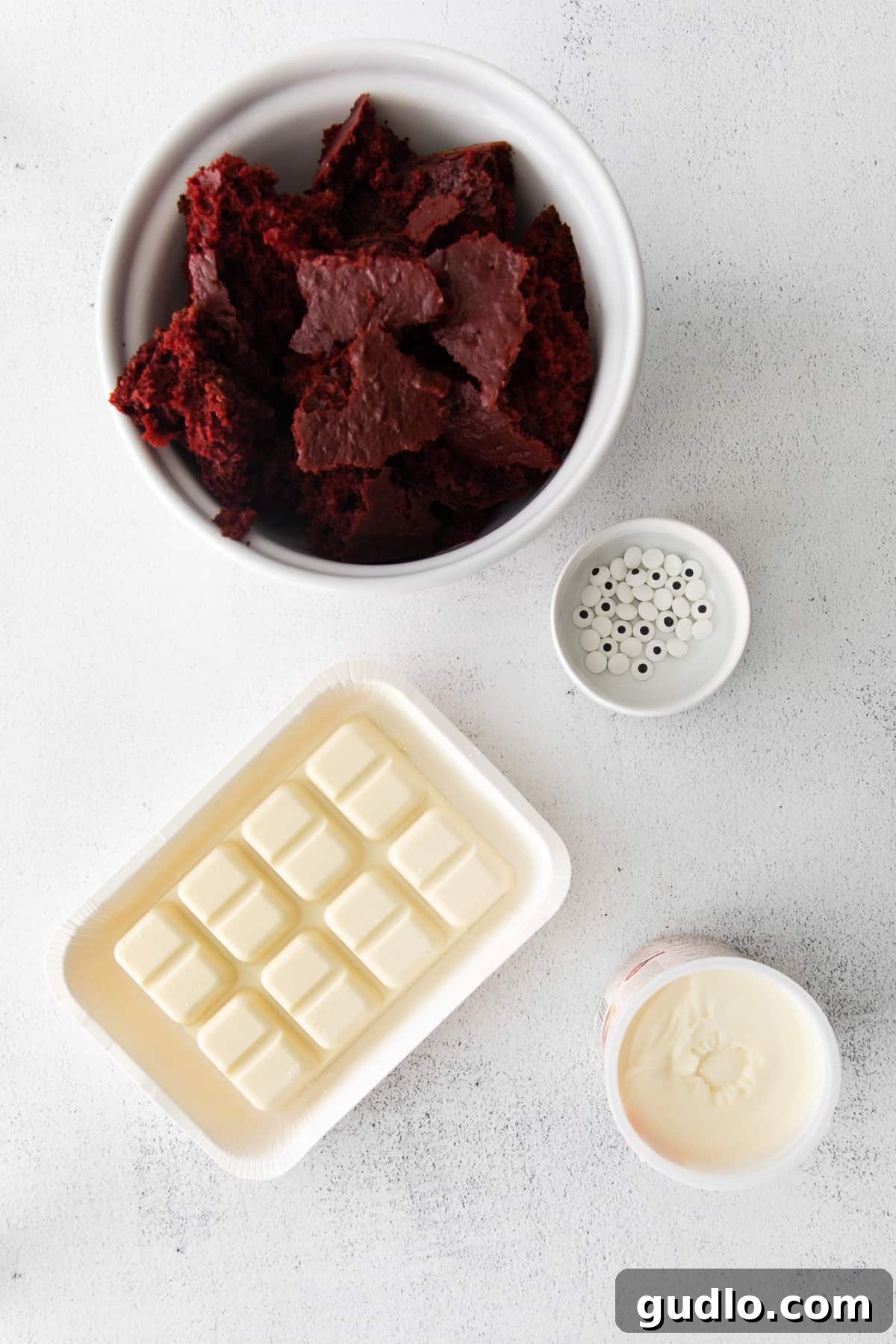Make these spooky Red Velvet Mummy Truffles for a fun Halloween treat the whole family can help make! A selection of raw ingredients including a red velvet cake mix box, cream cheese frosting, white chocolate bark, and candy eyes, laid out on a kitchen countertop.