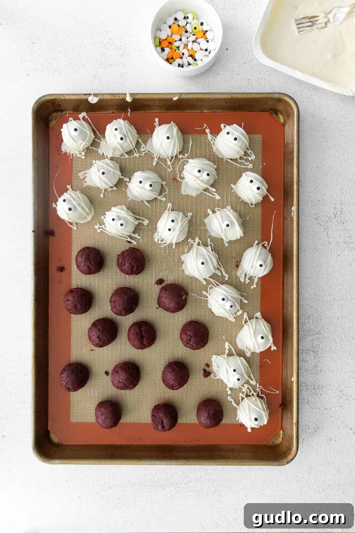 Red Velvet Mummy Truffles: Happy Halloween from these delicious dessert truffles! A baking sheet filled with red velvet truffles, some already dipped in white chocolate and adorned with candy eyes, demonstrating the decoration process.
