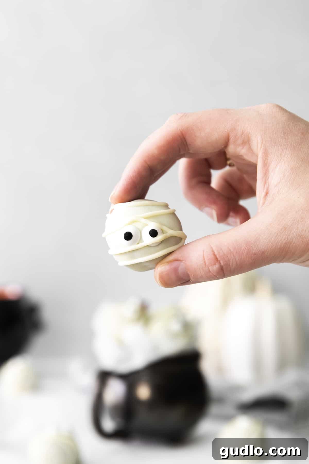 Red Velvet Mummy Truffles: Happy Halloween from these delicious dessert truffles! A hand gently holding a finished red velvet mummy truffle, beautifully decorated with candy eyes and delicate white chocolate 'bandages'.