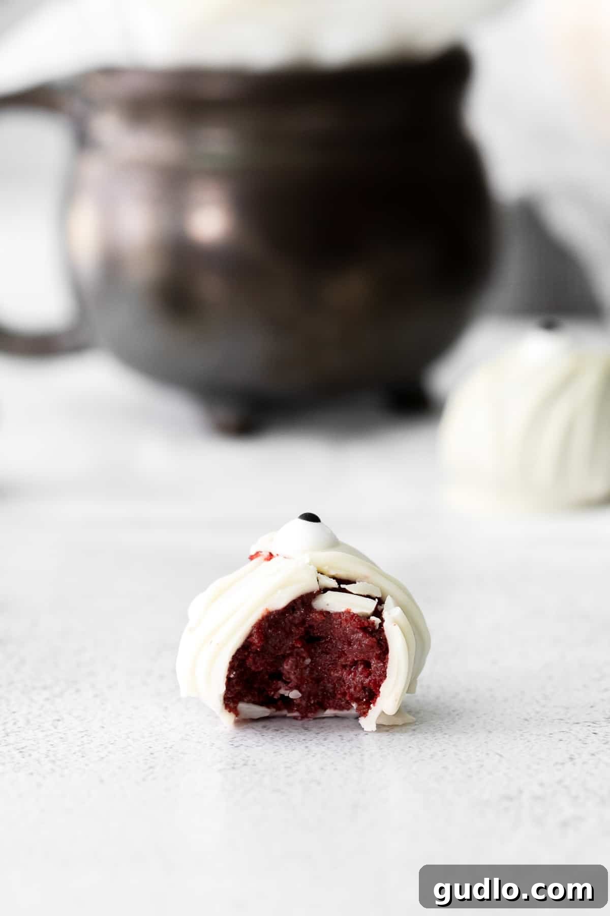 Red Velvet Mummy Truffles: Happy Halloween from these delicious dessert truffles! Close-up view of a red velvet mummy truffle with a bite taken out, revealing the moist, deep red cake interior beneath the white chocolate coating.
