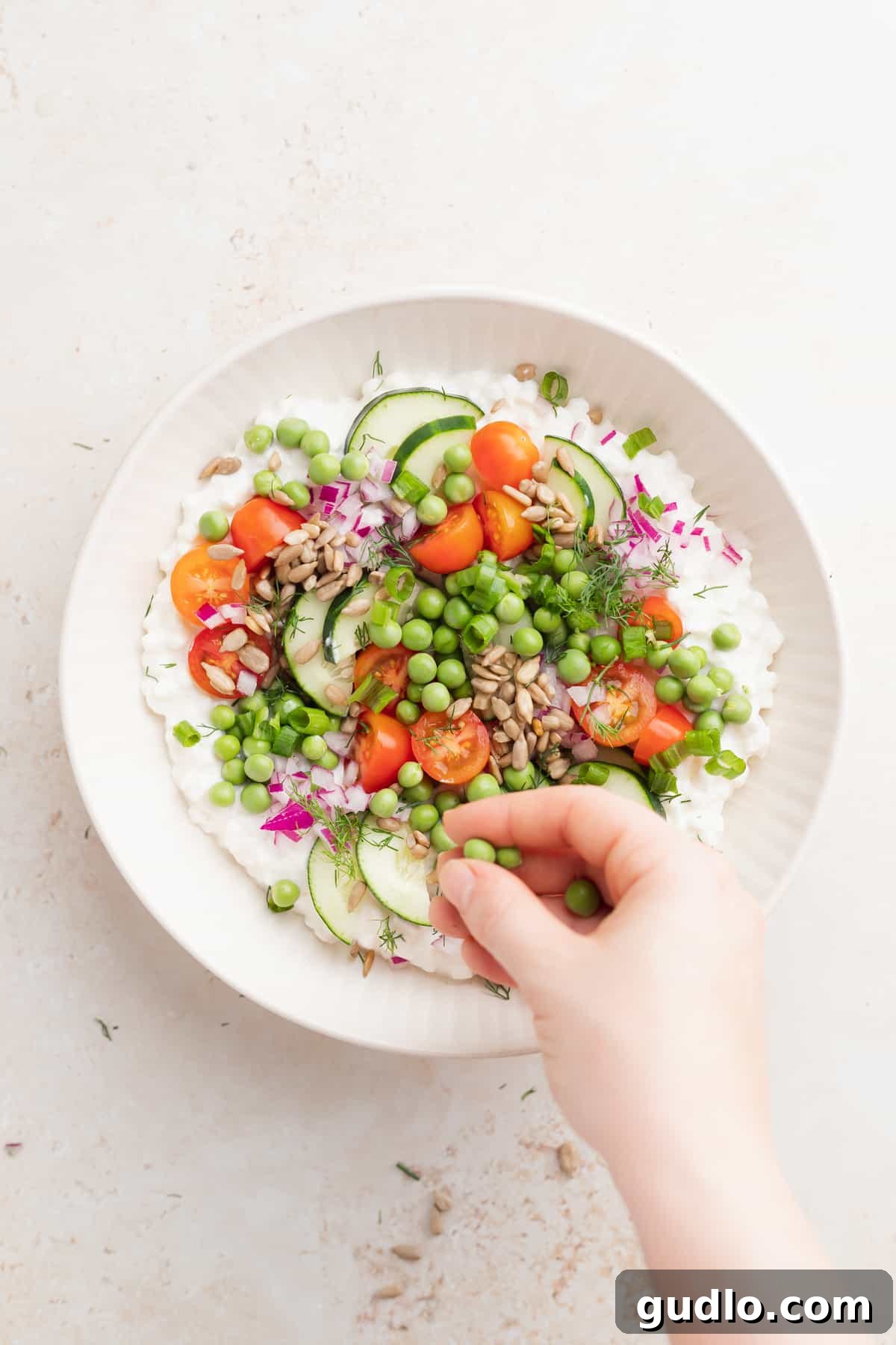 Adding Fresh English Peas to Cottage Cheese Salad A person's hand sprinkling fresh English peas over a bed of cottage cheese, which is already topped with other colorful vegetables, during the assembly of a healthy cottage cheese salad.