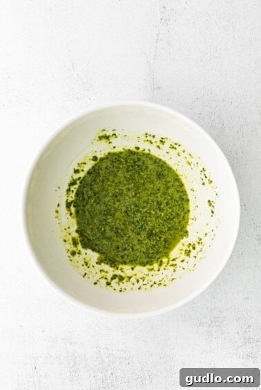 A bowl of vibrant green pesto being whisked with pasta water on a white surface, preparing for the pasta salad.