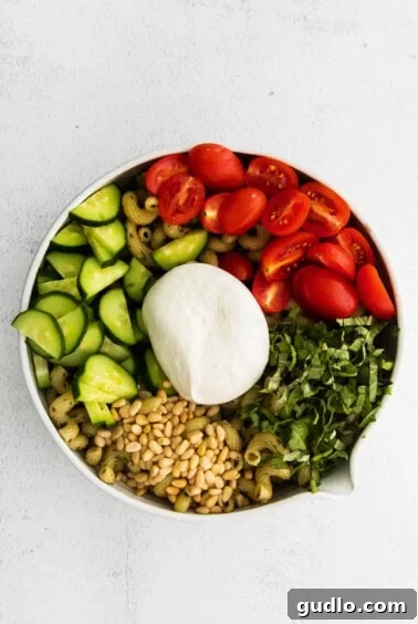 A large bowl containing pesto pasta salad with tomatoes, cucumbers, and other ingredients, ready for the burrata.