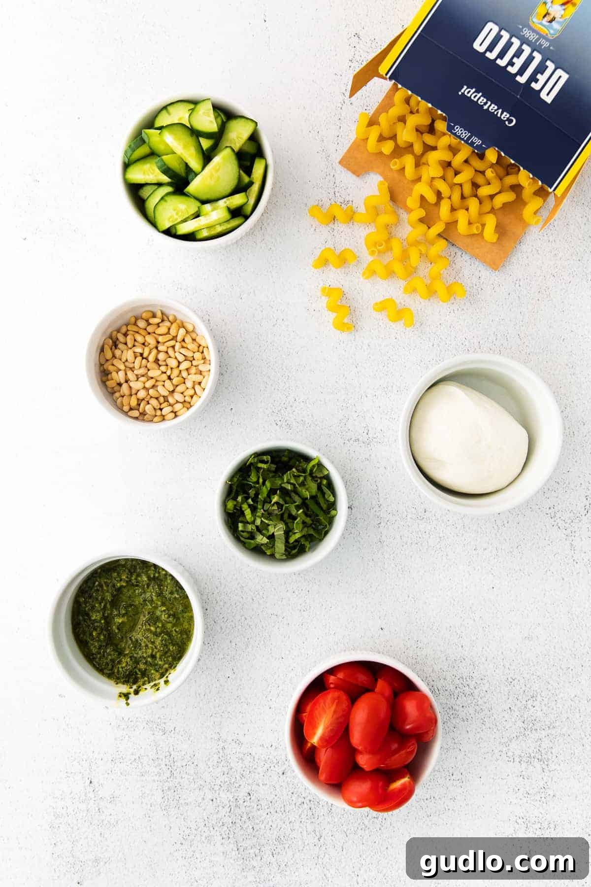 Key Ingredients for a Flavorful Pesto Pasta Salad A vibrant selection of fresh ingredients for pesto pasta salad laid out in separate bowls, including pasta, cherry tomatoes, cucumbers, and a jar of pesto.
