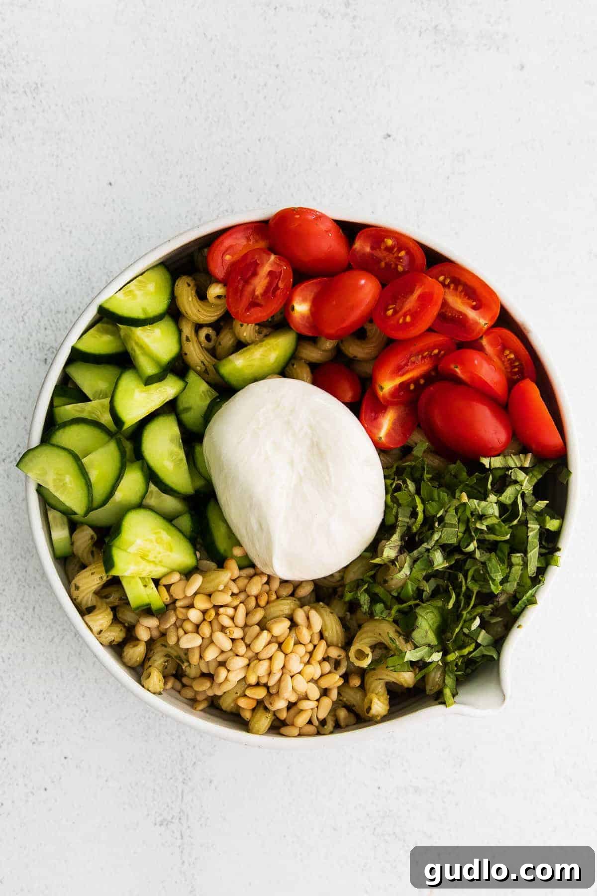 Pesto Pasta Salad Ready for Burrata Topping A close-up view of pesto pasta salad ingredients in a bowl, highlighted by a whole ball of burrata cheese resting on top.