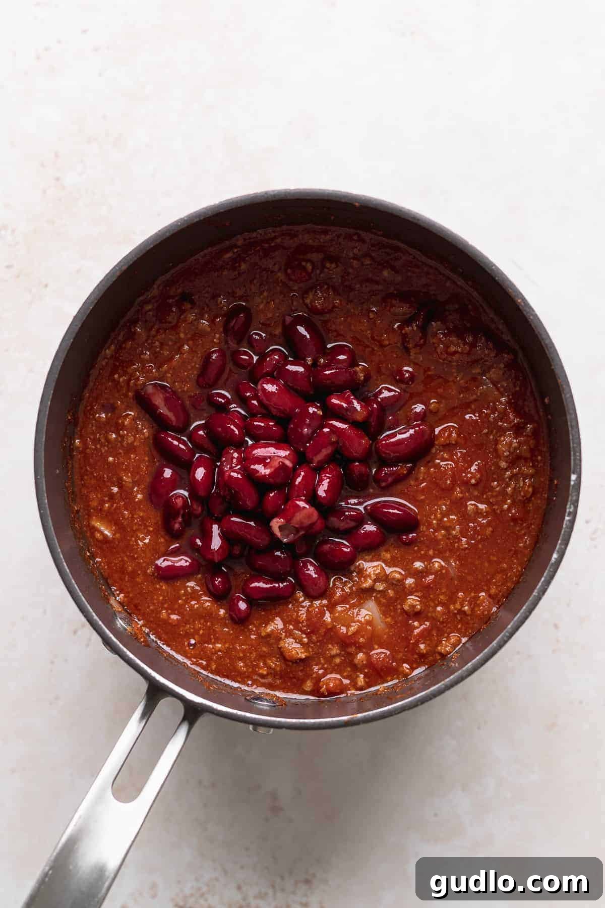 Ultimate Chili Cheese Fries 2 A hearty serving of chili beans ready to be spooned over crispy fries, presented in a pan on a white surface.