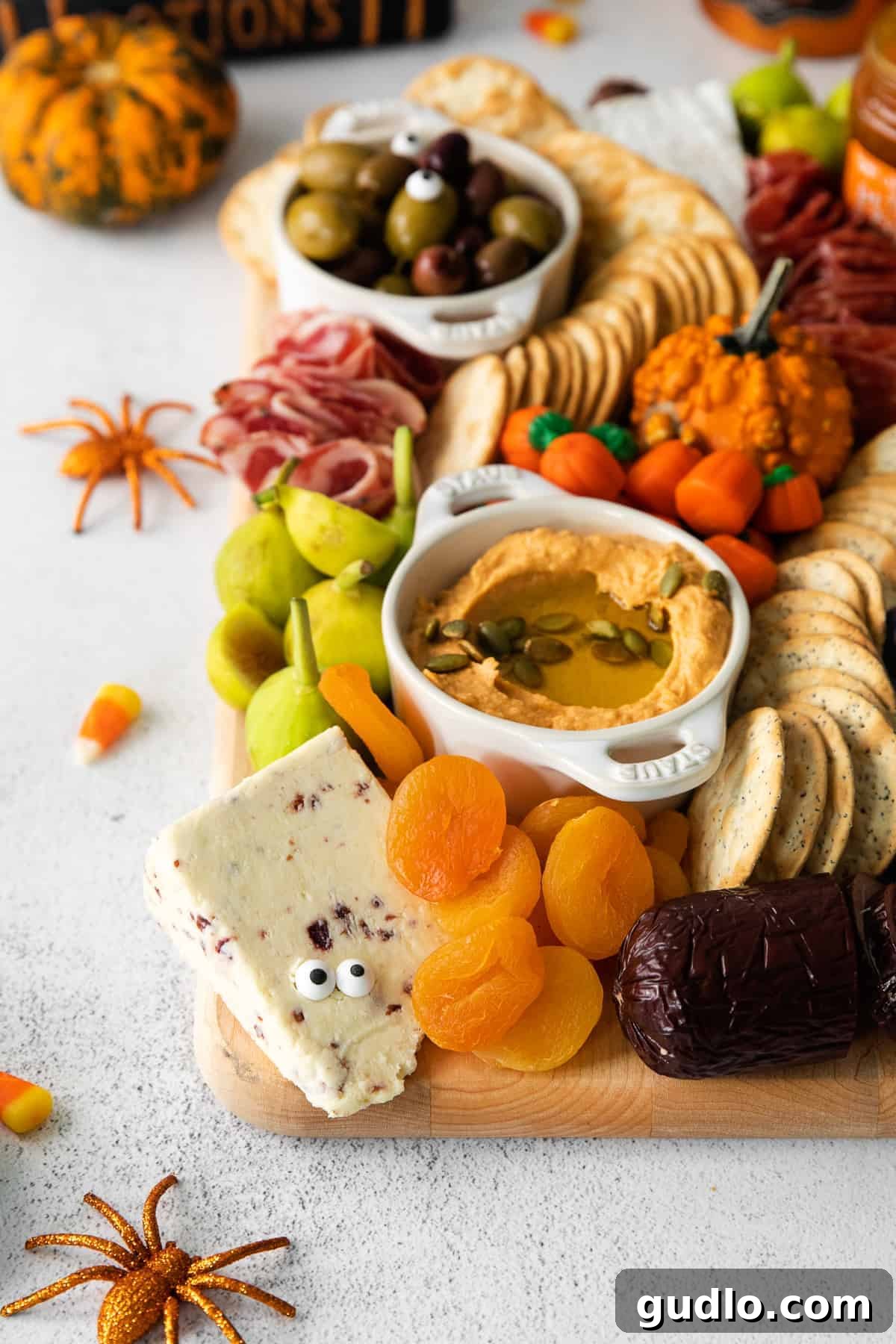 Ghoulish Grazing Board 5 A small bowl of dip garnished with sprigs of thyme, surrounded by cheese, crackers, and dark fruits on a Halloween board.