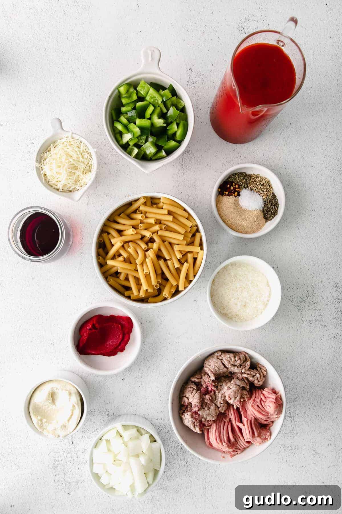 Ultimate Cheesy Ziti Bake 3 All essential ingredients for baked ziti, including meats, diced vegetables, spices, and cheeses, neatly arranged in small bowls on a kitchen countertop.