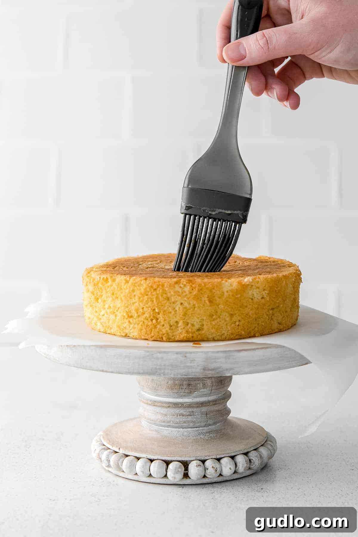 A silicone brush gently applying rich espresso to a layer of golden sponge cake, preparing it for the Tiramisu Layer Cake assembly.