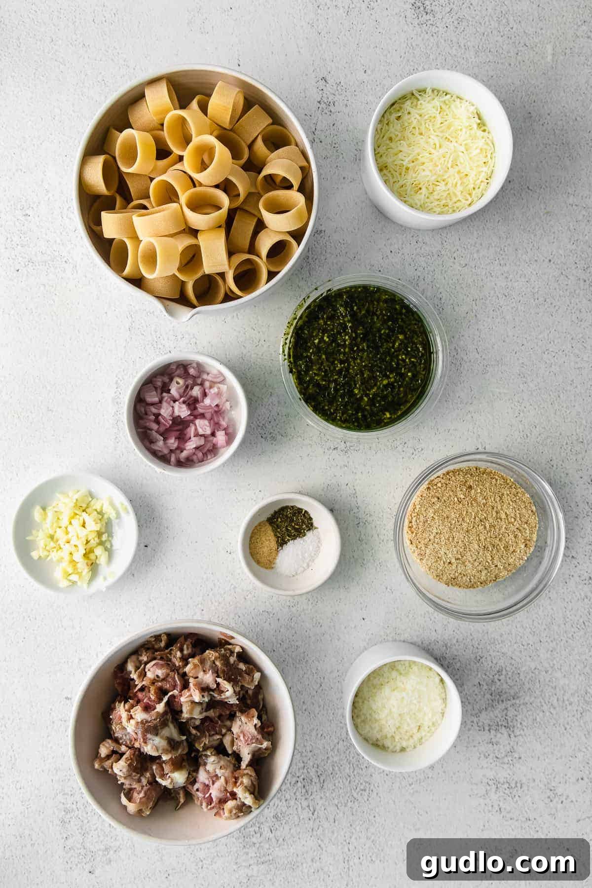 Fresh ingredients for pesto pasta including pasta, pesto, sausage, shallots, garlic, and cheeses laid out on a kitchen counter.