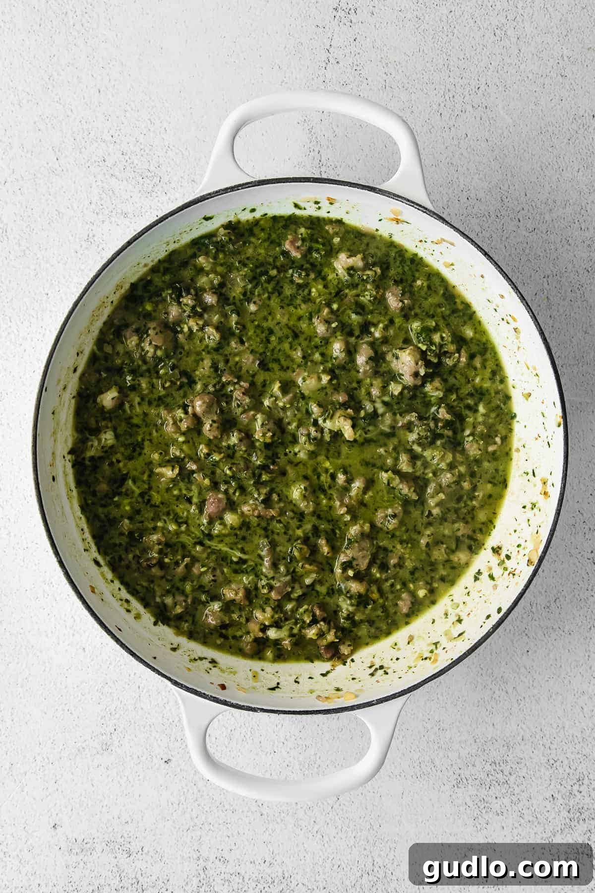 Rich green pesto sauce being stirred into cooked pasta and Italian sausage in a Dutch oven.