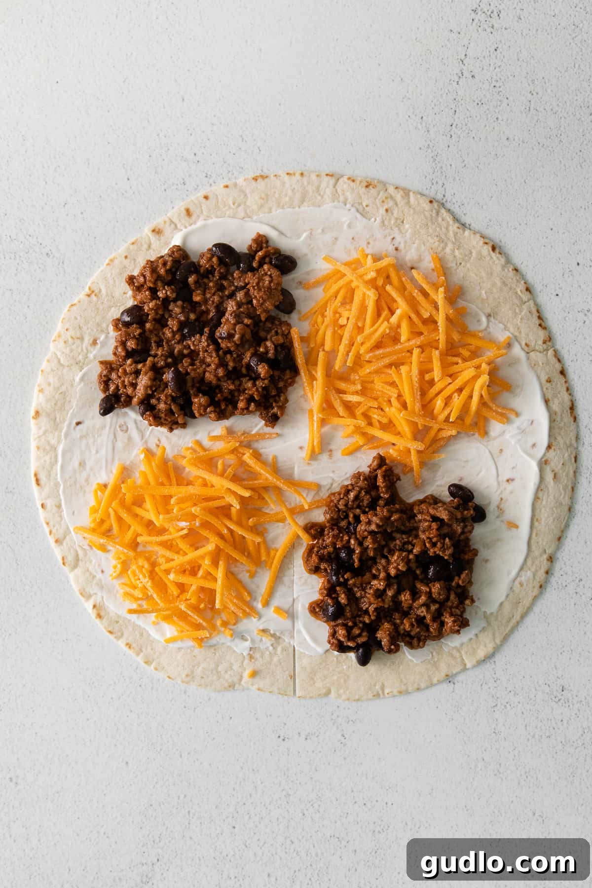 An XL flour tortilla neatly divided into four quadrants, with each section generously filled with taco-seasoned ground beef and shredded cheddar cheese, prepared for the unique folding technique.