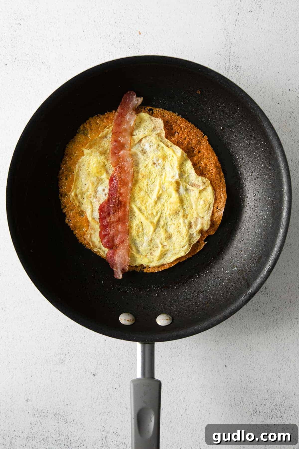 Assembling and Rolling Breakfast Taquitos The assembly process of a breakfast taquito in the skillet: a melted cheese wrap topped with an egg round, then a strip of bacon being carefully rolled into a tight taquito.