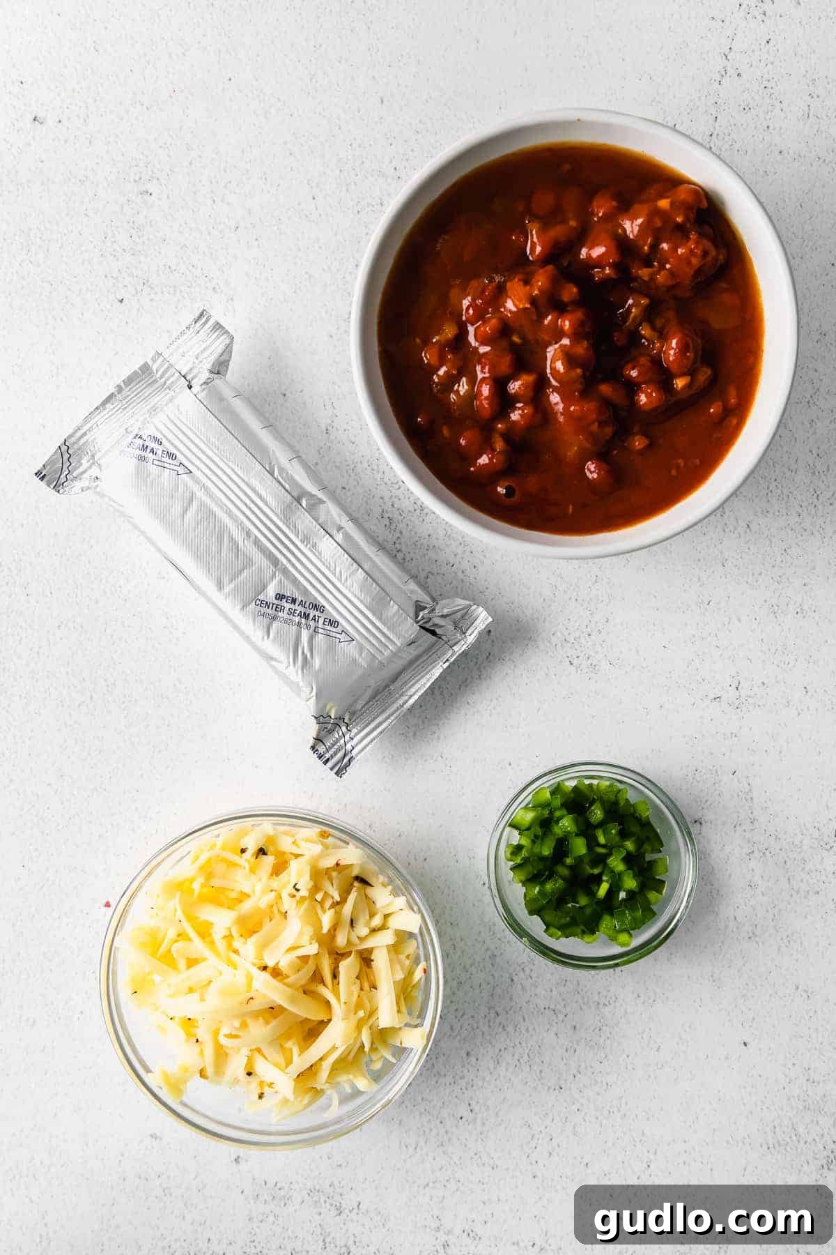 The four essential ingredients for chili cheese dip thoughtfully arranged on a wooden surface: a can of chili, a block of cream cheese, a pile of freshly shredded pepper jack cheese, and a single vibrant green jalapeño.