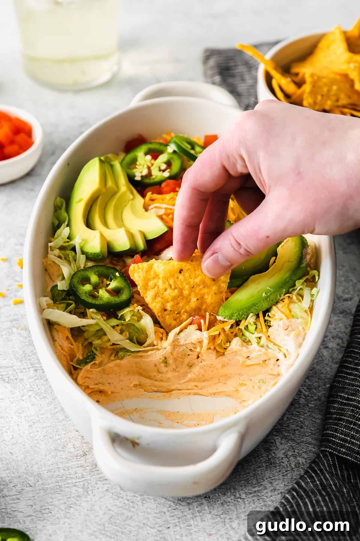 A crisp tortilla chip being dipped into a bowl of rich, layered taco dip, showcasing its creamy texture and fresh toppings.