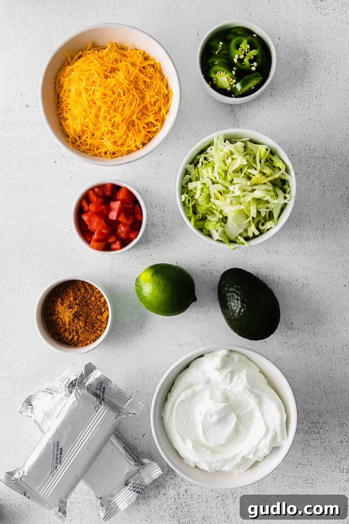 Individual bowls showcasing the fresh, vibrant ingredients for taco dip, including cream cheese, Greek yogurt, tomatoes, avocado, and jalapeños.