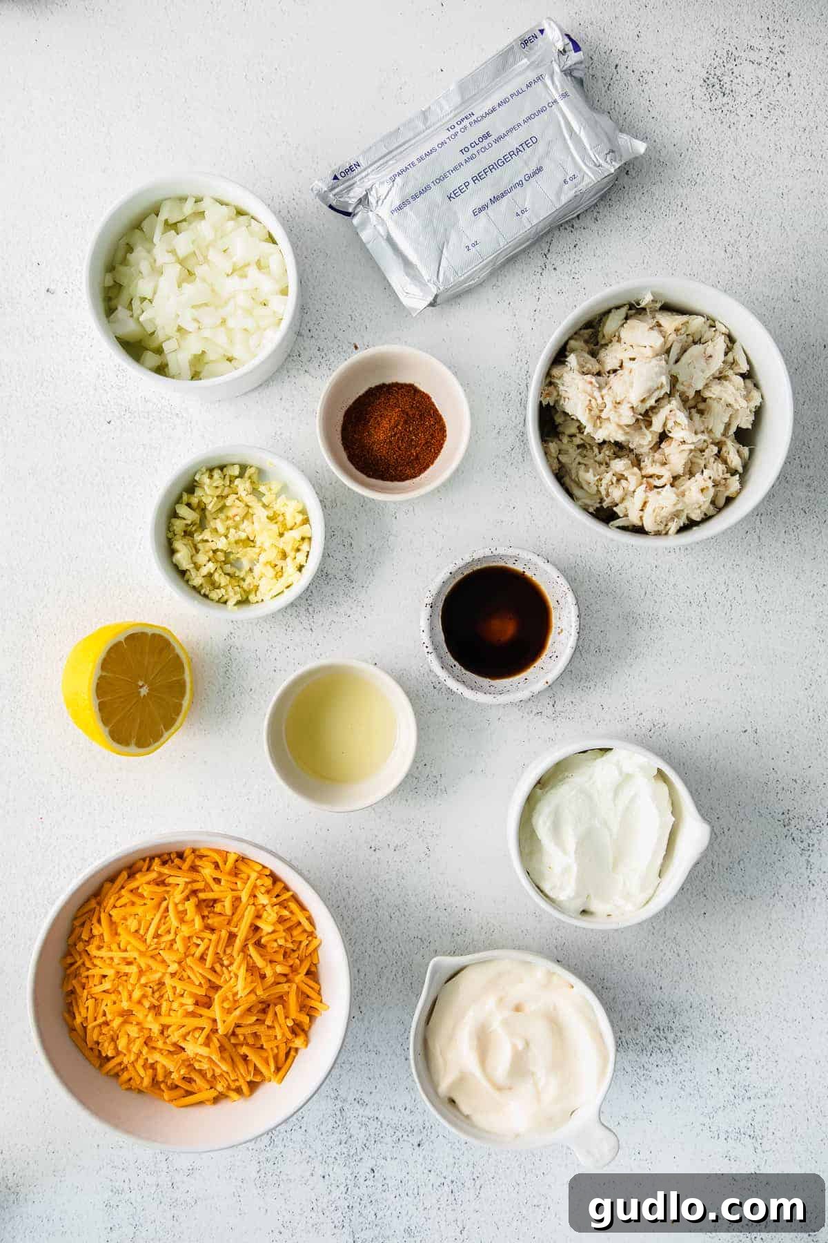 Ultimate Cheesy Crab Dip 4 All ingredients for hot crab dip laid out on a kitchen counter: sour cream, mayonnaise, cream cheese, fresh garlic, onion, spices, and a container of lump crab meat.