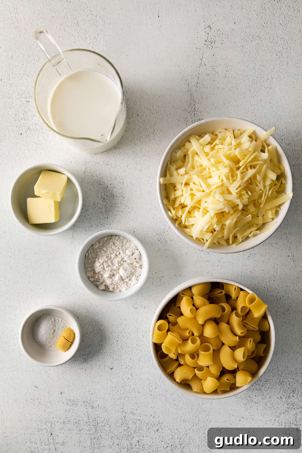 A perfectly arranged selection of fresh ingredients on a light countertop, including pasta, butter, flour, milk, white cheddar cheese, salt, and ground mustard, ready for cooking.