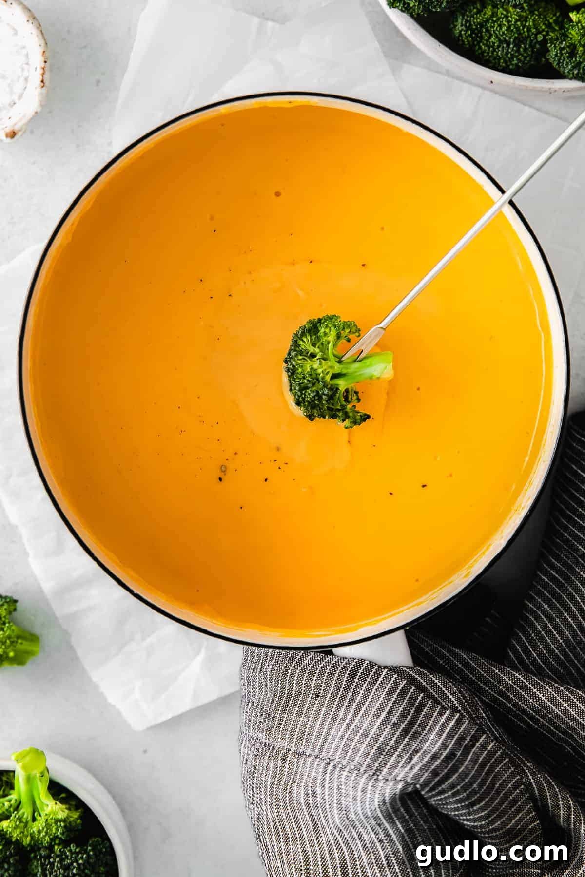 A hand dipping a vibrant green broccoli floret into a bowl of rich, yellow cheese sauce.