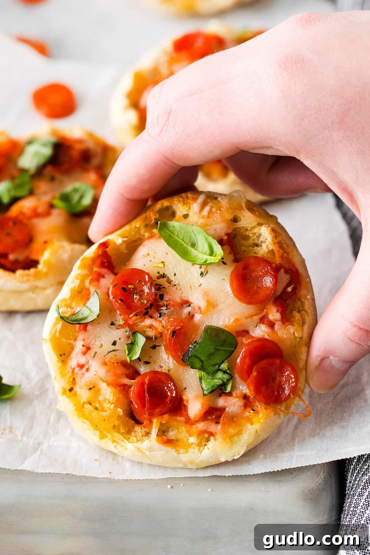 Let dinner be a breeze with English Muffin Pizzas! This kid-friendly dinner idea is made with toasted English muffins and your favorite pizza toppings. A hand holding a freshly baked English muffin pizza, showcasing melted cheese and pepperoni.