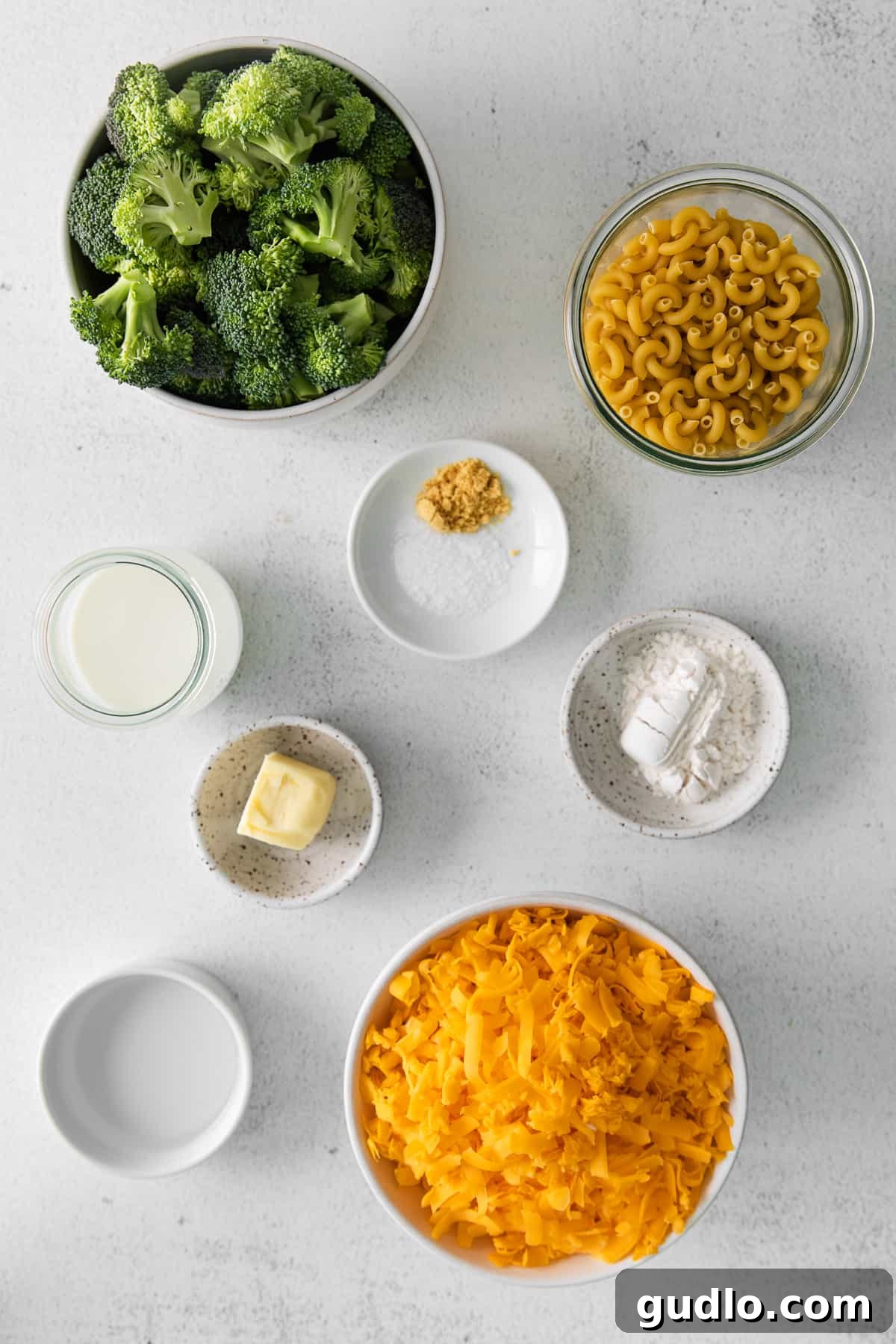 Broccoli Mac and Cheese Ingredients Fresh ingredients laid out on a kitchen countertop, including macaroni noodles, broccoli florets, blocks of cheddar cheese, butter, milk, and spices, ready for cooking.